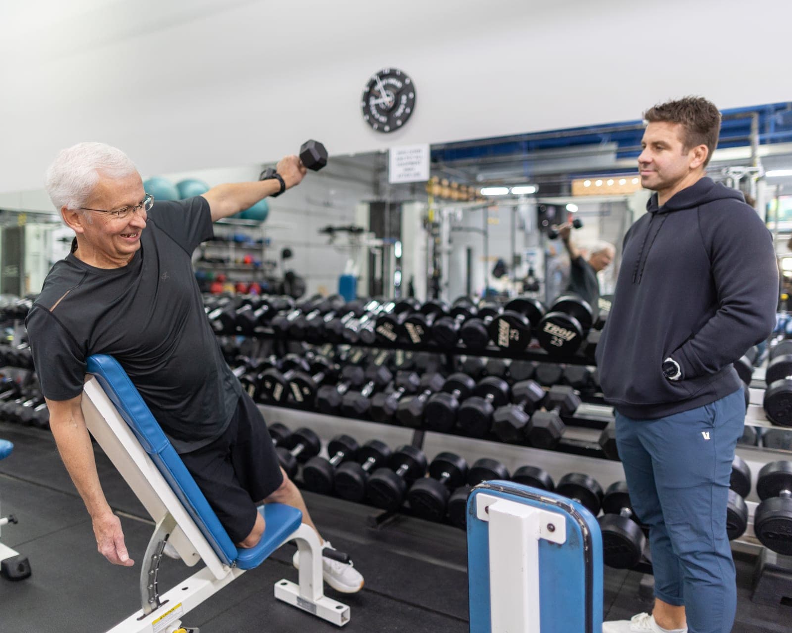 A white-haired CNE member laughing mid lateral-raise on an incline bench while his trainer stands beside him, with the dumbbell racks behind them