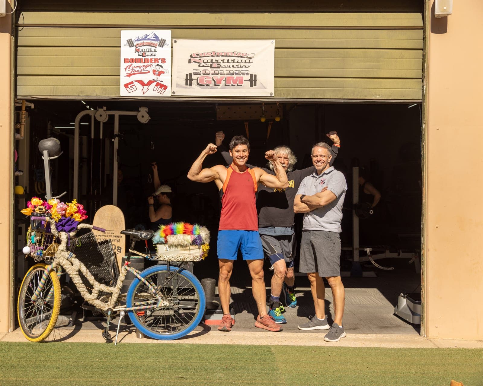 Three people laughing in CNE's open roll-up doorway beneath the Customized Nutrition & Exercise Boulder Gym banner: a member flexing, a white-haired member flexing beside him, and owner Rick Jones with his arms folded