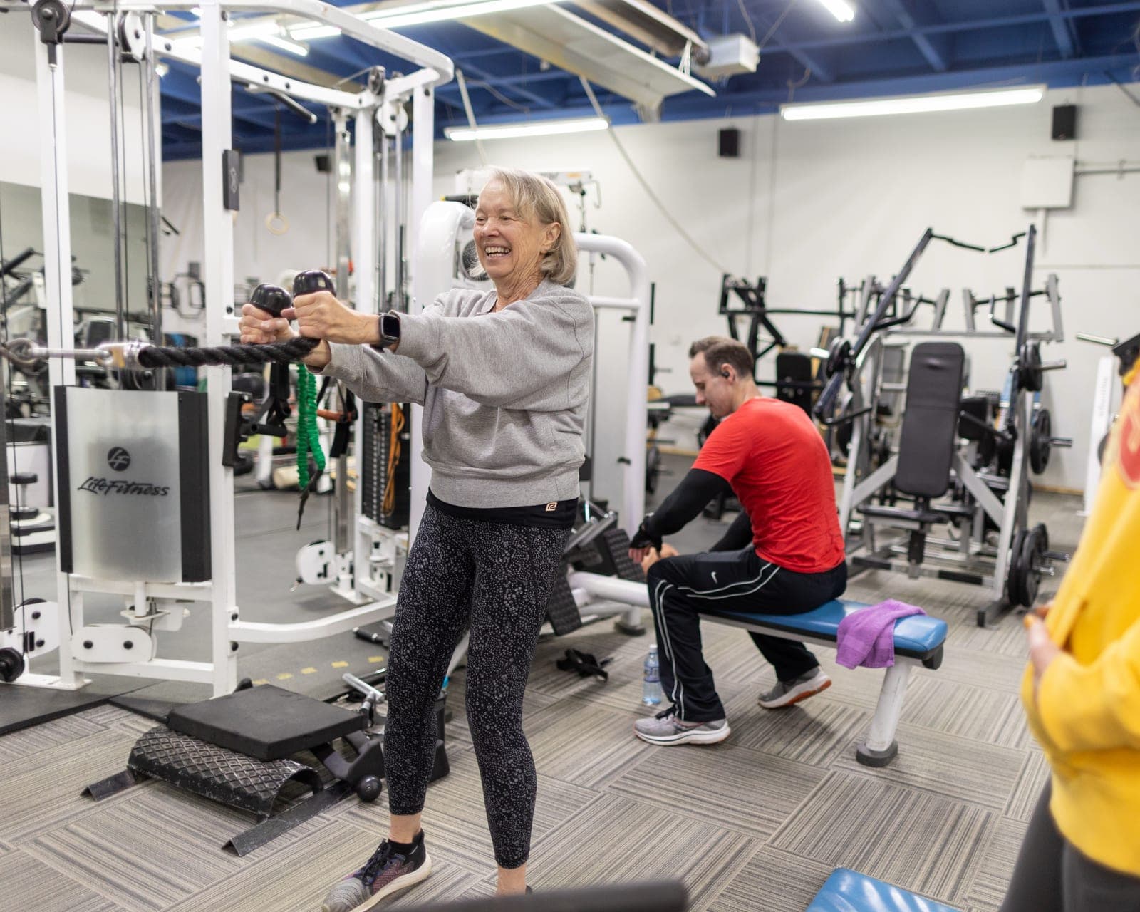 An older CNE member grinning as she presses a cable rope out in front of her at the functional trainer, with another member training on a bench behind