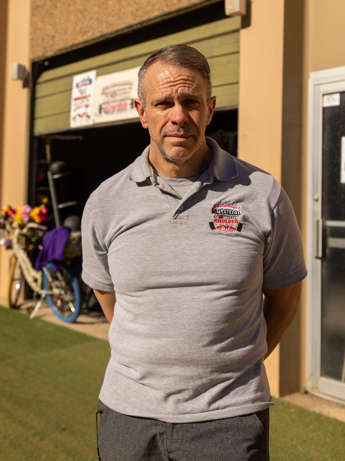 Rick Jones, owner of CNE, standing outside the gym's open roll-up door in a Customized Nutrition & Exercise polo shirt