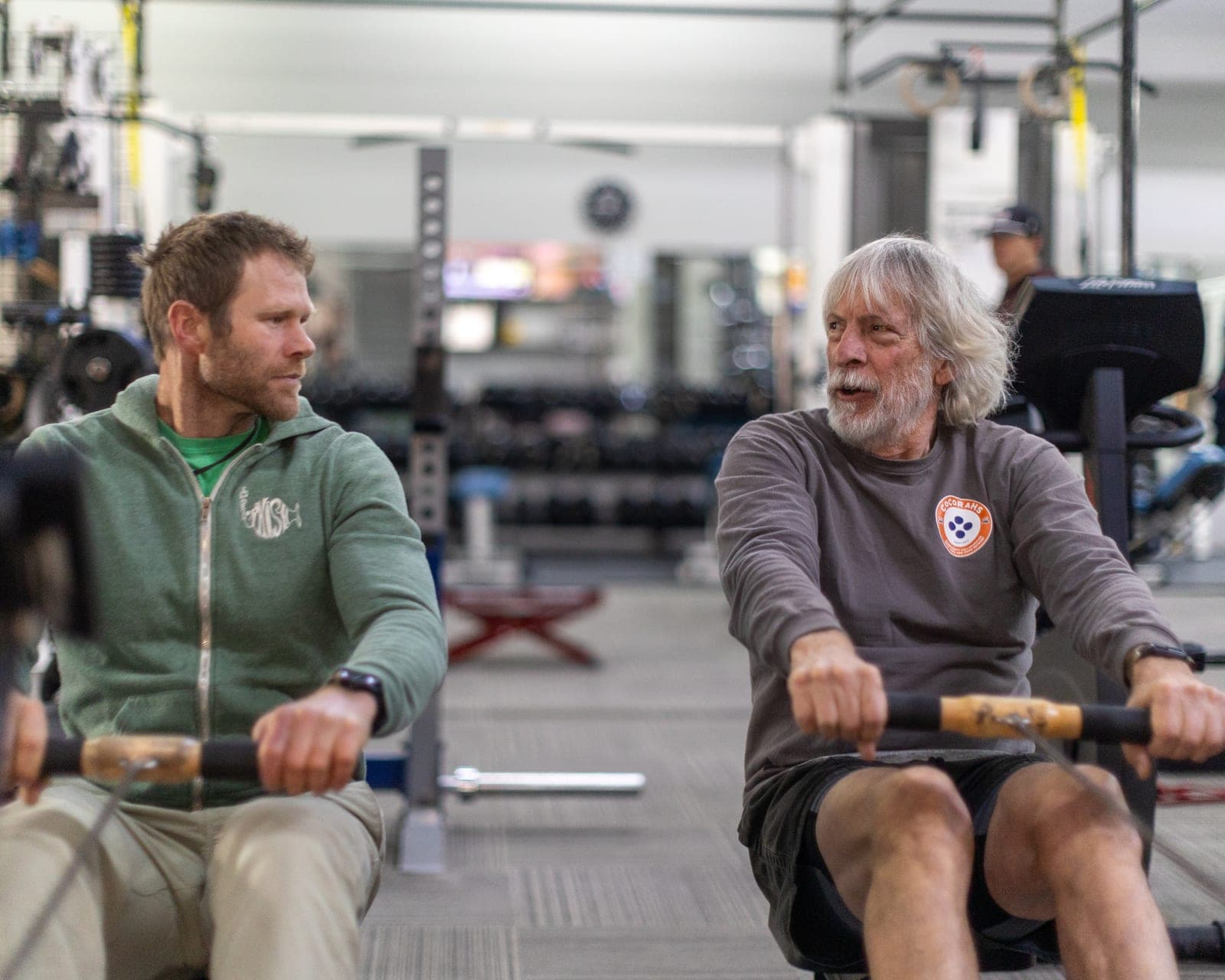 Two CNE members mid-stroke on side-by-side rowing machines, mid-conversation — one in his thirties, one white-haired and bearded